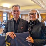 Ernst Uhing und Andrea Gebhard bei der Verabschiedung von Uhing vom Vorstand der BAK. Ein Mann und eine Frau lächeln in die Kamera, im Hintergrund sitzen Personen auf Stühlen an einem Tisch.