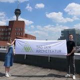 Eine junge Frau und ein junger Mann halten die Fahne zum Tag der Architektur hoch. Im Hintergrund sind die Gehry-Bauten und der Fernsehturm in Düsseldorf zu sehen.