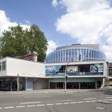 Stadttheater Münster Ein Gebäude an einer Straßer gebaut, im Hintergrund sind Bäume und ein blauer Himmel mit Wolken.